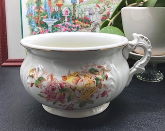 May include: A white ceramic chamber pot with a gold rim and ornate handle. The pot is decorated with a floral pattern featuring pink, yellow, and orange roses. The pot has a wide opening and a pedestal base.