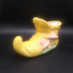 May include: A yellow ceramic shoe planter with pink and green mushroom accents. The planter is shaped like a shoe with a pointed toe and a curved heel.
