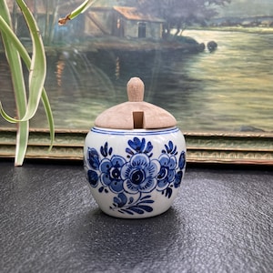 May include: A small white ceramic jar with a blue and white floral design. The jar has a wooden lid with a small opening in the centre.