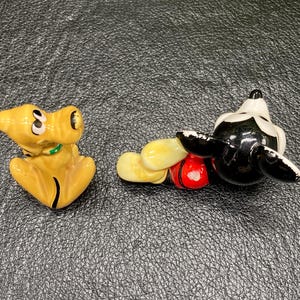 May include: A ceramic figurine of Mickey Mouse in a red and yellow outfit, lying on his side with his head turned to the side.  A ceramic figurine of Pluto, a yellow dog, is standing on all fours with his head turned to the side.
