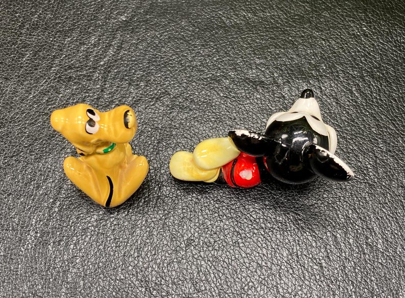 May include: A ceramic figurine of Mickey Mouse in a red and yellow outfit, lying on his side with his head turned to the side.  A ceramic figurine of Pluto, a yellow dog, is standing on all fours with his head turned to the side.