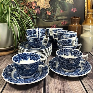 May include: A collection of blue and white china teacups and saucers. The cups and saucers feature a detailed landscape design. The teacups are stacked in groups, with some arranged on a wooden surface. A vintage floral painting is in the background.