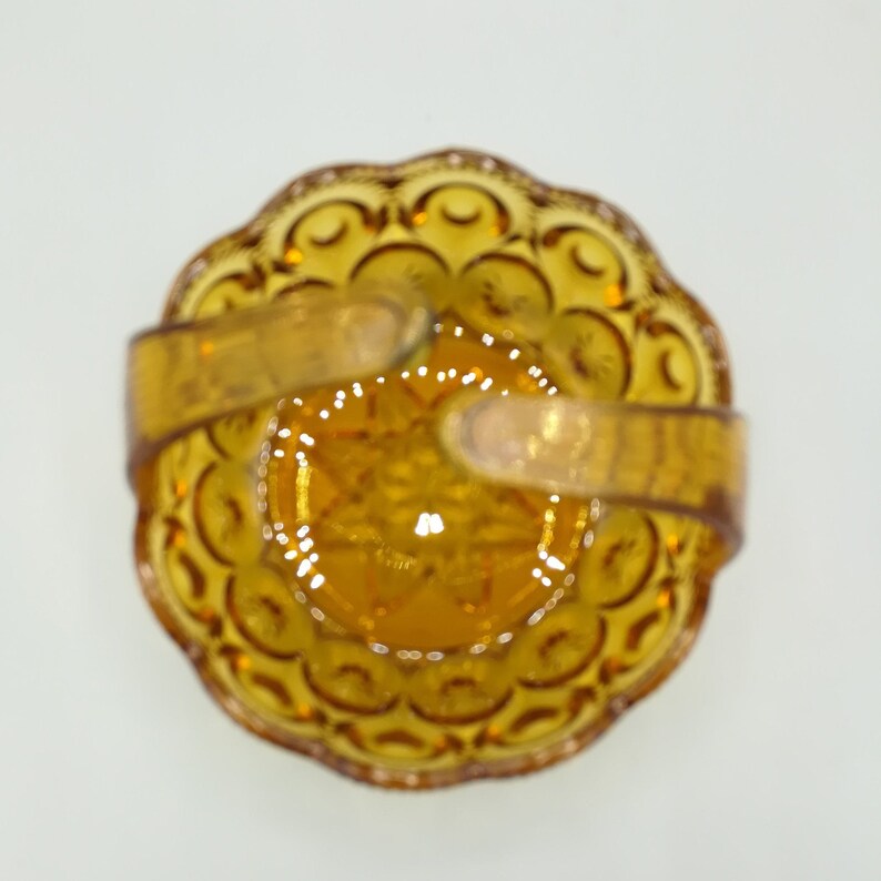 May include: An overhead view of a vintage amber-colored glass basket. The basket has a scalloped edge and a starburst pattern in the center. It features a handle made of the same amber glass material.