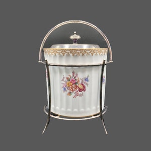 May include: A white porcelain ice bucket with a floral design and a silver lid and stand. The bucket has a gold trim around the top and a floral design on the side.
