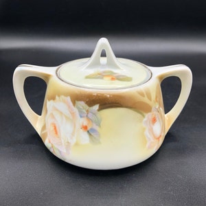 May include: A white porcelain sugar bowl with a lid, featuring a hand-painted design of roses and leaves in shades of pink, green, and brown. The bowl has two handles and a gold trim around the rim.