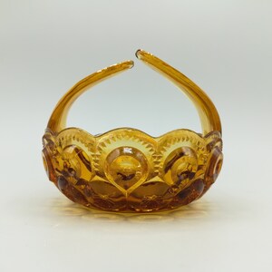 May include: An amber-colored glass basket with a handle. The basket has a scalloped rim and is decorated with a repeating pattern of circles and starbursts. The handle arches upwards from the sides of the basket.
