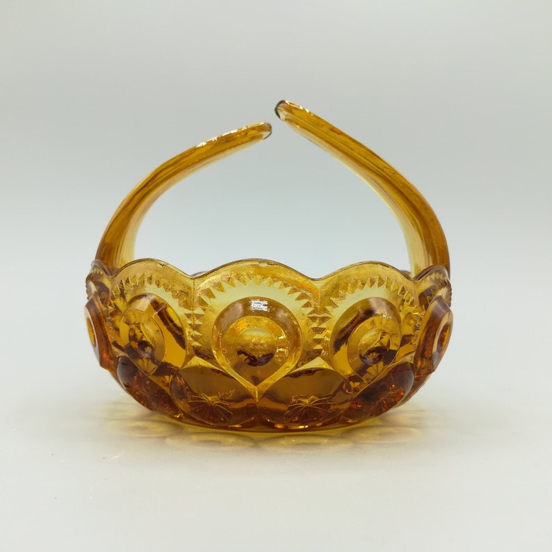 May include: An amber-colored glass basket with a handle. The basket has a scalloped rim and is decorated with a repeating pattern of circles and starbursts. The handle arches upwards from the sides of the basket.