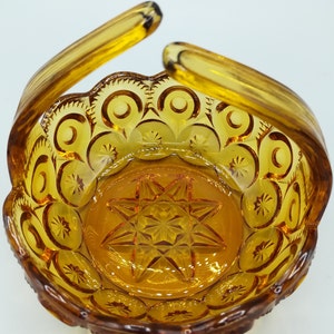 May include: Amber-colored glass basket with a handle. The basket features a starburst design in the center and a repeating pattern of crescent moons and stars around the rim. The handles curve upwards, creating a decorative and functional piece.
