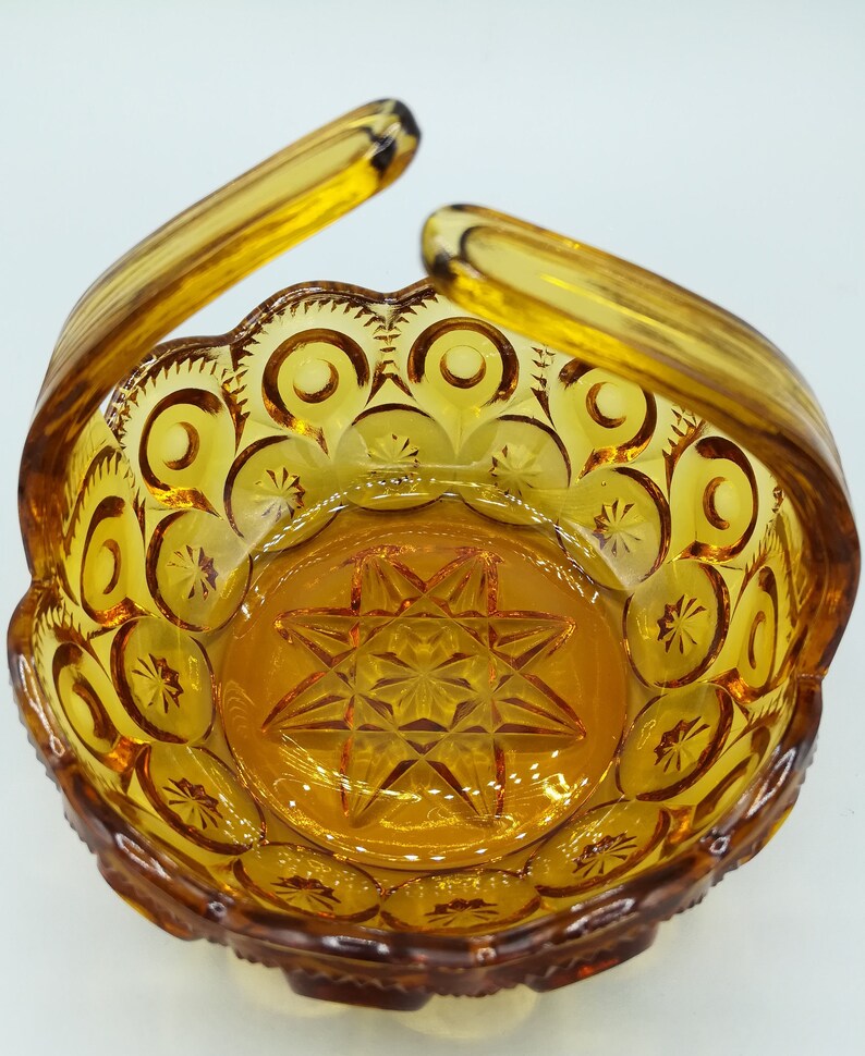 May include: Amber-colored glass basket with a handle. The basket features a starburst design in the center and a repeating pattern of crescent moons and stars around the rim. The handles curve upwards, creating a decorative and functional piece.