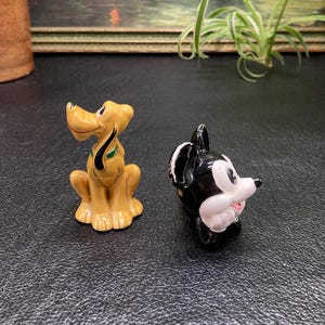 May include: A pair of ceramic figurines, one of Pluto the dog and one of Mickey Mouse. Pluto is a yellow dog with a green collar and Mickey Mouse is black with white gloves and a red mouth.