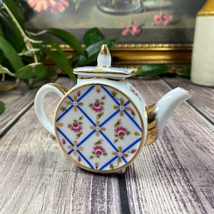 May include: A small white porcelain teapot featuring a blue lattice pattern with gold accents and pink rose floral motifs. The teapot has a round body, a matching lid with a pointed finial, a curved handle, and a short spout with gold trim.