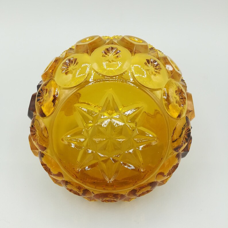 May include: A round, amber-colored glass bowl with a starburst pattern in the center. The bowl has a textured surface with raised circular and geometric designs around the edges. The glass has a warm, golden hue.