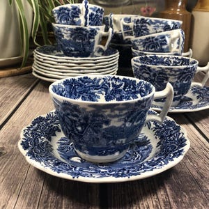 May include: A set of white ceramic teacups and saucers featuring a blue transferware pattern of rural landscapes and foliage. Each cup has a scalloped rim and a curved handle, while the saucers display a matching intricate blue floral border.