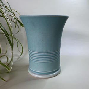 May include: A teal ceramic vase with a flared top and a circular base. The vase features a series of horizontal, white-painted lines around the middle. The vase is approximately 8 inches tall. A houseplant with long, thin leaves is visible in the background.
