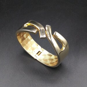 May include: A gold-toned metal bangle bracelet with a unique, abstract design. The bracelet is hinged and has a textured finish.