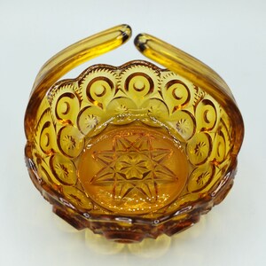 May include: Amber-colored glass basket with a handle. The basket features a starburst pattern on the base and a scalloped rim with crescent moon and star designs. The handle arches upward from the sides.