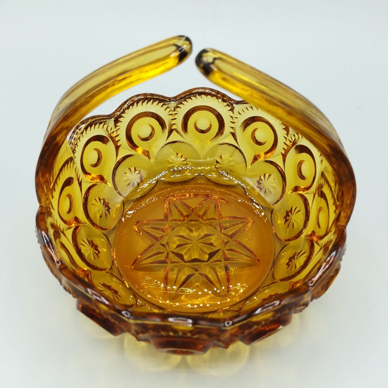 May include: Amber-colored glass basket with a handle. The basket features a starburst pattern on the base and a scalloped rim with crescent moon and star designs. The handle arches upward from the sides.