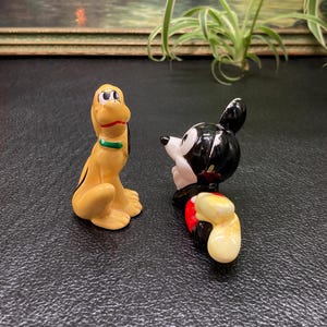 May include: A ceramic figurine of Mickey Mouse in a red and yellow outfit, looking to the left, and Pluto, a yellow dog with a green collar, looking to the right.