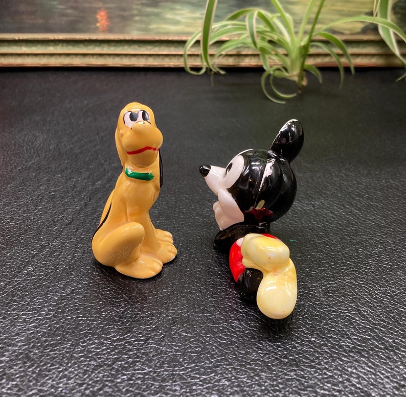 May include: A ceramic figurine of Mickey Mouse in a red and yellow outfit, looking to the left, and Pluto, a yellow dog with a green collar, looking to the right.