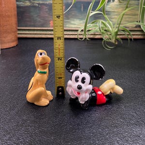 May include: A ceramic figurine set featuring Mickey Mouse and Pluto. Mickey is lying on his stomach with his arms crossed and Pluto is standing on all fours. Both figurines are painted in their signature colors.