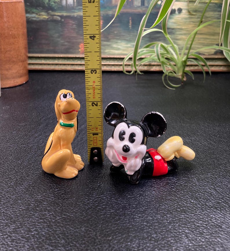 May include: A ceramic figurine set featuring Mickey Mouse and Pluto. Mickey is lying on his stomach with his arms crossed and Pluto is standing on all fours. Both figurines are painted in their signature colors.