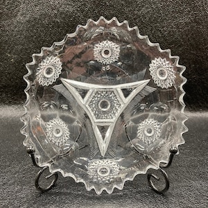 May include: A clear glass plate with a scalloped edge and a geometric design in the center. The plate has a starburst pattern around the edge and a central design of a triangle with a starburst in the center.