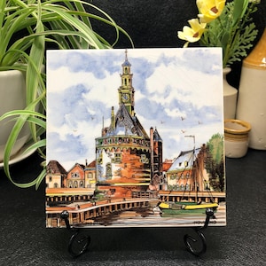 May include: A vintage ceramic tile depicting a watercolor painting of a Dutch town with a tall tower and a canal. The tile is white with blue and brown accents and has the word "Holland" printed on the bottom.