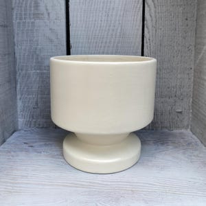 May include: A solid off-white ceramic planter with a smooth, matte finish. The vessel features a cylindrical upper body that tapers inward towards a wide, circular pedestal base, creating a stable and balanced structure for holding plants.