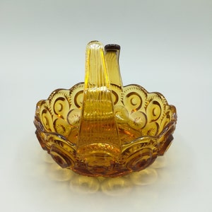 May include: Amber-colored glass basket with a handle. The basket has a scalloped edge with a repeating pattern of crescent moons and stars. The handle is vertical and has a ribbed design. The glass has a glossy finish.
