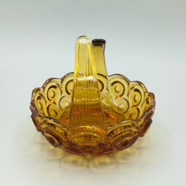 May include: Amber-colored glass basket with a handle. The basket has a scalloped edge with a repeating pattern of crescent moons and stars. The handle is vertical and has a ribbed design. The glass has a glossy finish.