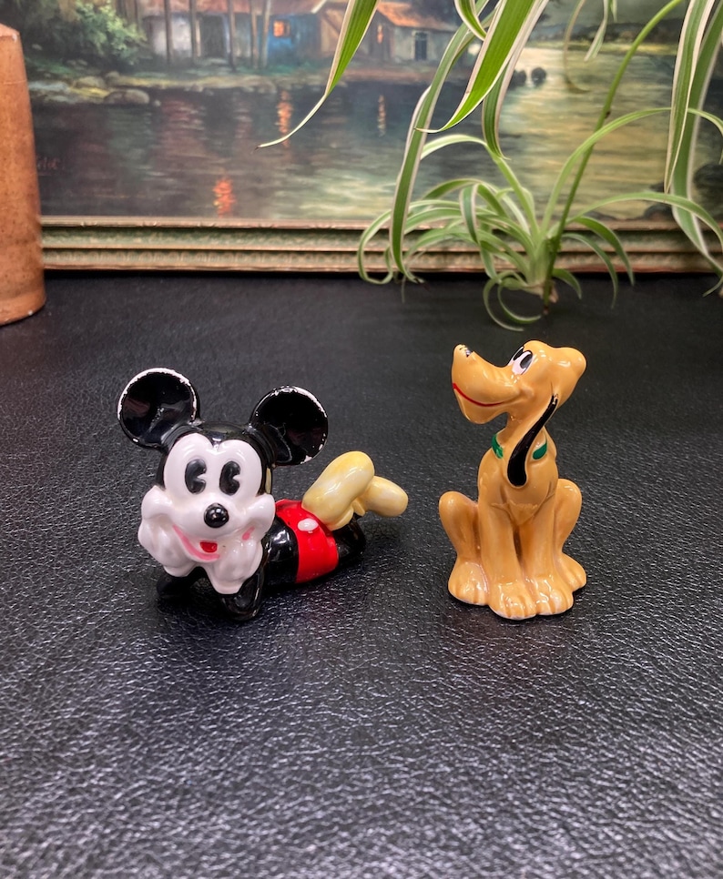 May include: A vintage ceramic figurine set featuring Mickey Mouse and Pluto. Mickey is lying down with his signature red shorts and white gloves. Pluto is standing with his tail curled. Both figurines are painted in bright colors.