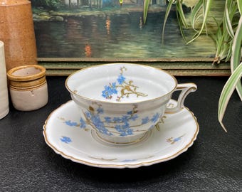 May include: A white porcelain teacup and saucer with a delicate floral pattern. The cup and saucer are decorated with blue forget-me-not flowers and gold accents.