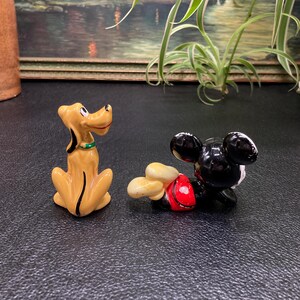 May include: A ceramic figurine of Pluto the dog, sitting with his tail curled, and a ceramic figurine of Mickey Mouse lying on his side with his arms outstretched.