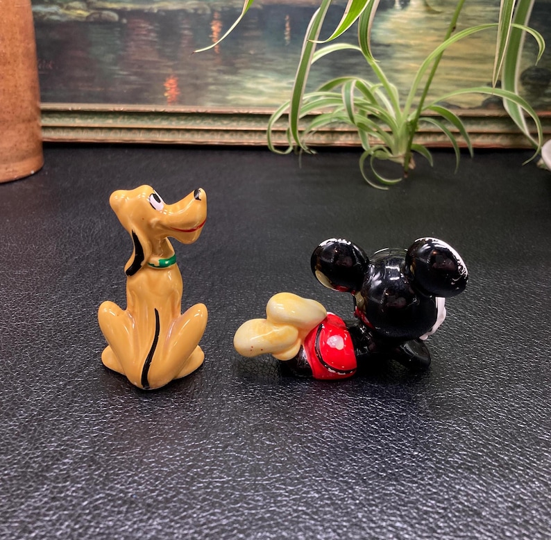 May include: A ceramic figurine of Pluto the dog, sitting with his tail curled, and a ceramic figurine of Mickey Mouse lying on his side with his arms outstretched.
