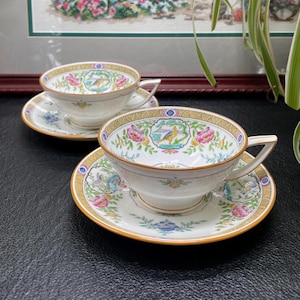 May include: Two white teacups and saucers with a decorative floral and bird design. The cups and saucers have a gold trim and are set on a dark surface. The teacups have a handle.