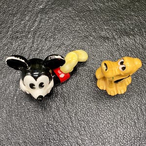 May include: A black and white ceramic figurine of Mickey Mouse with red shorts and a yellow tail. A yellow ceramic figurine of Pluto the dog is also pictured.