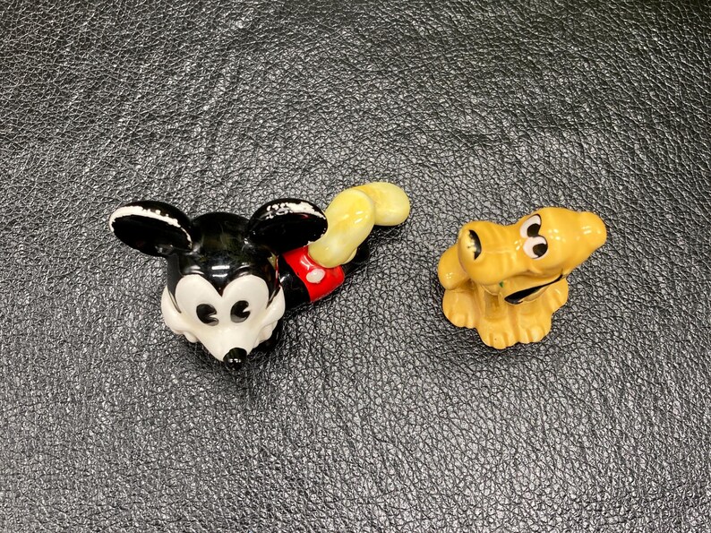 May include: A black and white ceramic figurine of Mickey Mouse with red shorts and a yellow tail. A yellow ceramic figurine of Pluto the dog is also pictured.
