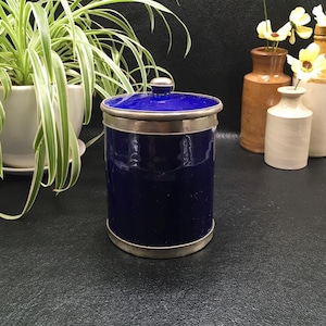 Peut inclure: Un pot cylindrique bleu cobalt avec un couvercle et une garniture argentée. Le pot a un petit bouton rond sur le dessus du couvercle. Le pot est posé sur une surface noire, avec des plantes et des vases en arrière-plan.