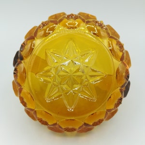 May include: Amber-colored glass bowl with a starburst design in the center. The bowl has a circular shape with a textured, scalloped rim. The glass is transparent, allowing light to pass through, and has a warm, golden hue.