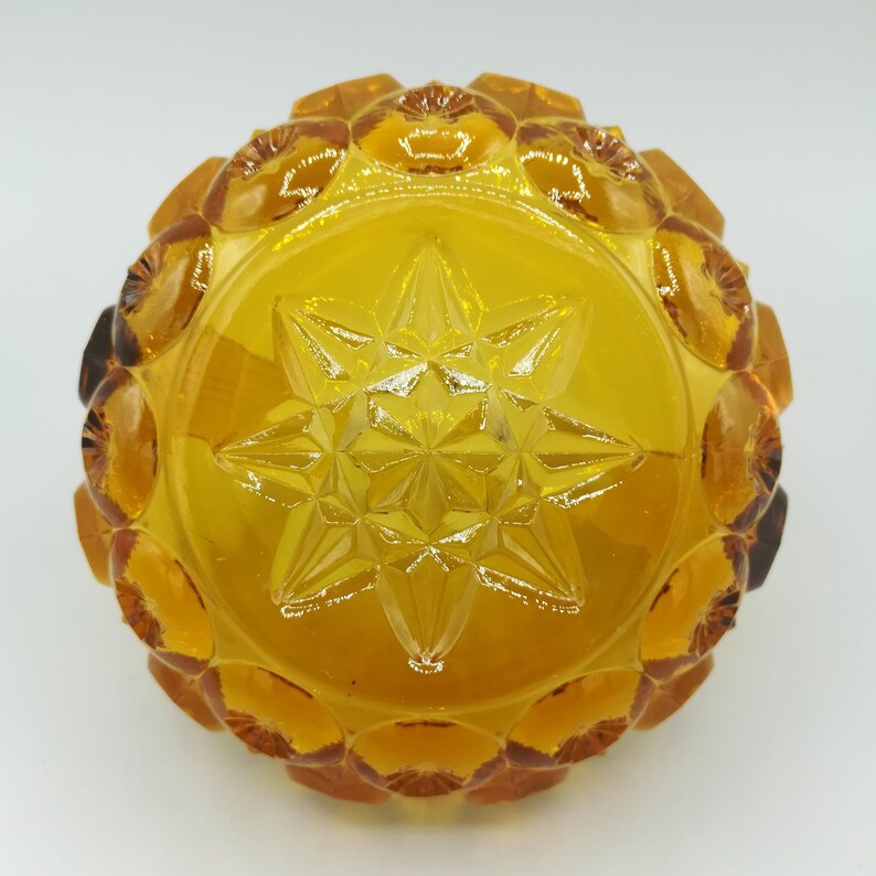 May include: Amber-colored glass bowl with a starburst design in the center. The bowl has a circular shape with a textured, scalloped rim. The glass is transparent, allowing light to pass through, and has a warm, golden hue.
