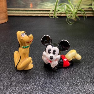 May include: A ceramic figurine of Mickey Mouse wearing a red and white outfit, lying on his side with his head resting on his hand.  A ceramic figurine of Pluto, a yellow dog with a green collar, is standing upright.