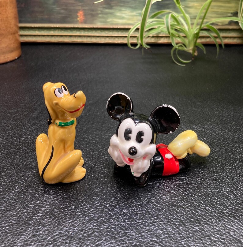 May include: A ceramic figurine of Mickey Mouse wearing a red and white outfit, lying on his side with his head resting on his hand.  A ceramic figurine of Pluto, a yellow dog with a green collar, is standing upright.