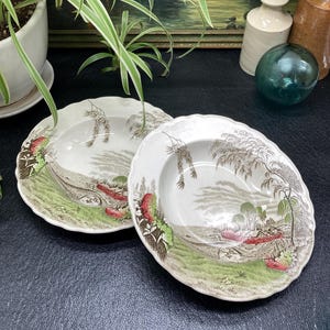 May include: Two white ceramic soup bowls with a green and brown landscape design. The bowls have a scalloped edge and a raised rim.