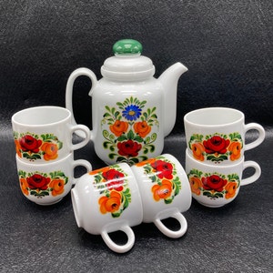 May include: A white teapot and six teacups with a floral design featuring red, orange, and blue flowers. The teapot has a green lid and a spout. The cups have a white base and a floral design around the top.