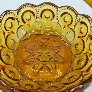 May include: Amber-colored glass basket with a handle. The basket features a starburst pattern in the center and a scalloped rim with circular and star-shaped designs. The glass has a glossy finish, reflecting light.