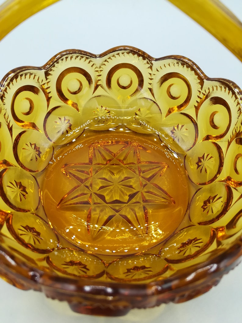 May include: Amber-colored glass basket with a handle. The basket features a starburst pattern in the center and a scalloped rim with circular and star-shaped designs. The glass has a glossy finish, reflecting light.