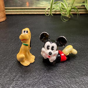 May include: A ceramic figurine of Mickey Mouse lying down with his head resting on his hand. He is wearing red shorts with white polka dots. Next to him is a ceramic figurine of Pluto, a yellow dog with black ears and a green collar.