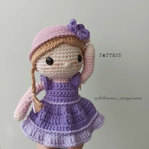 Puede incluir: Muñeca de crochet con ropa rosa y morada. La muñeca tiene cabello castaño, un sombrero rosa con una flor morada y ojos marrones. La muñeca se está sosteniendo la mano en la cabeza. El texto "PATTERN @philomena_amigurumis" es visible en la imagen.