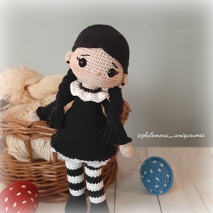 May include: A crocheted doll with black hair, a black dress, white and black striped leggings, and black shoes. The doll is holding a small blue and white crocheted ball. The doll is sitting on a wicker basket with yarn inside. The text "@philomena_amigurumis" is visible in the lower right corner of the image.