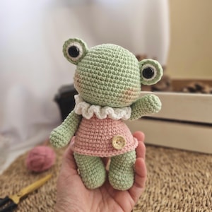 Puede incluir: Una muñeca de rana de ganchillo hecha a mano en verde claro y rosa. La rana tiene ojos negros grandes, un cuello blanco con volantes y un vestido rosa con un botón de madera. La muñeca se sostiene en una mano.